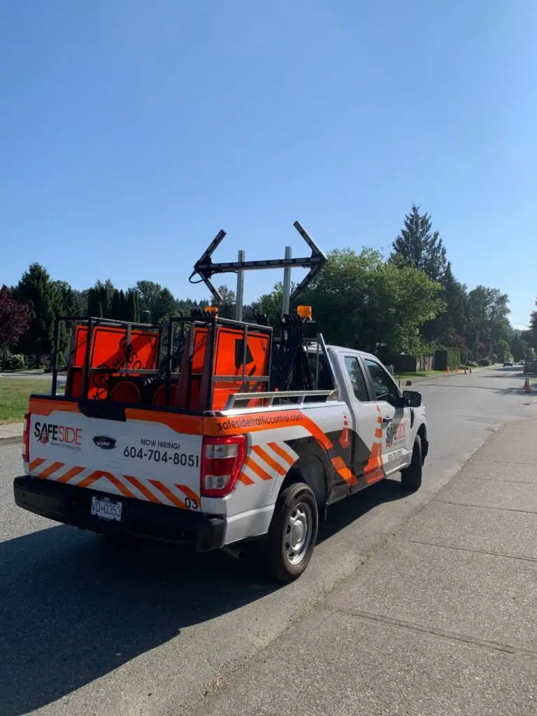 A white pickup truck with orange safety equipment and Traffic Control Services signs in the back is parked on a residential street on a sunny day.