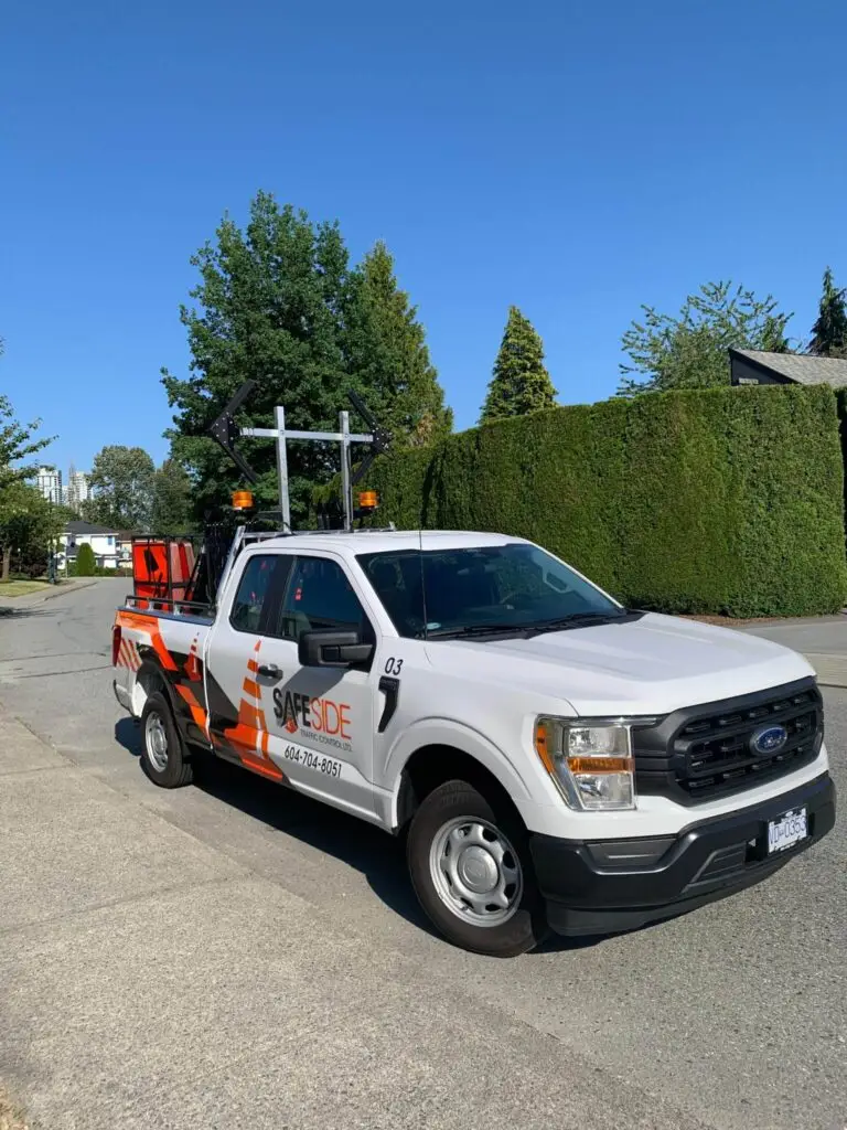 A white SafeSide company pickup truck with orange markings and Traffic Control Services equipment in the bed is parked on a residential street on a sunny day.