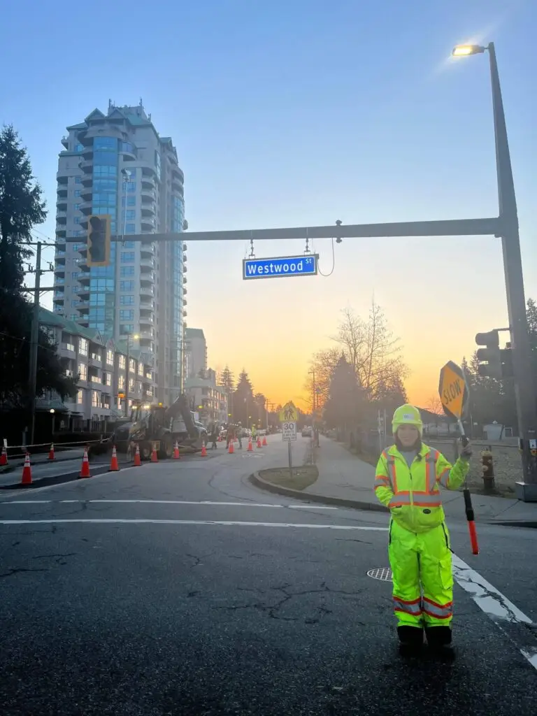 A construction worker in high-visibility clothing holds a "SLOW" sign at an intersection on Westwood Street, providing Traffic Control Services amid traffic cones and buildings in the background at dusk.